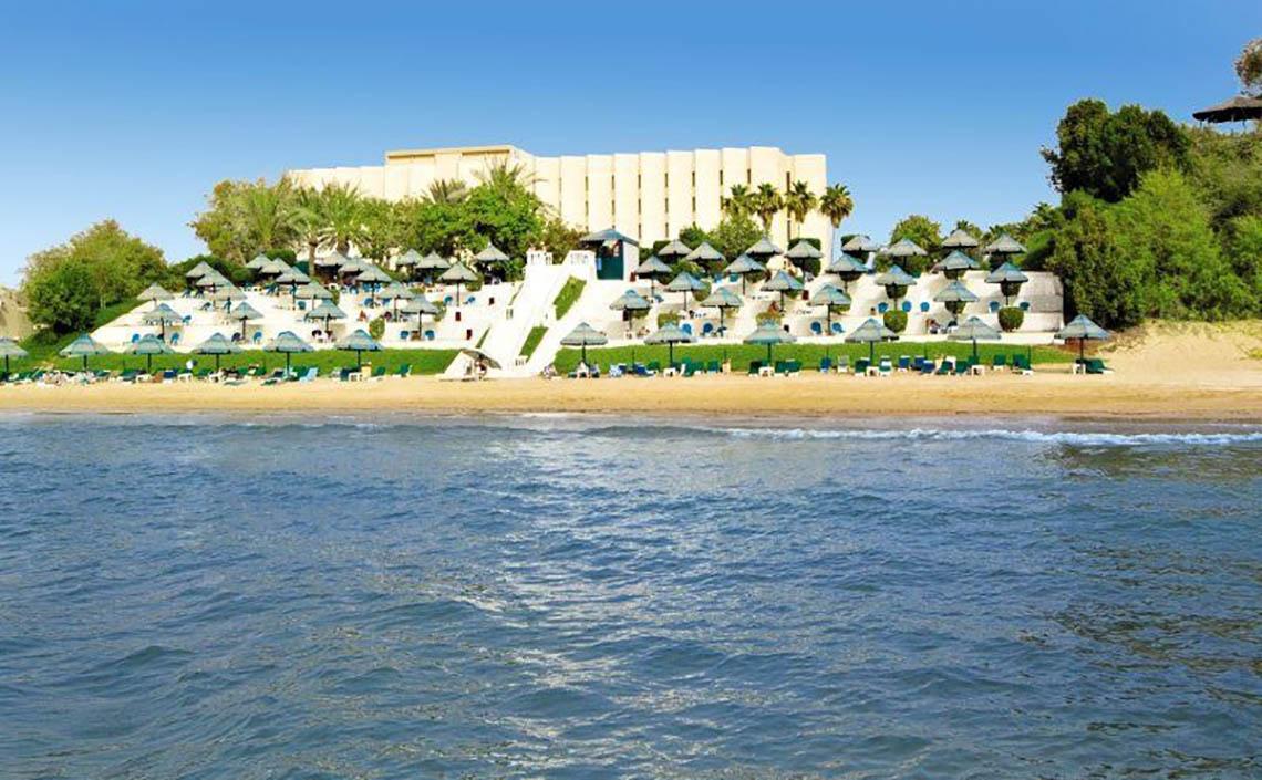 Außenansicht des BM Beach Hotel mit mehreren Sonnenschirmen und Liegestühlen am Strand.