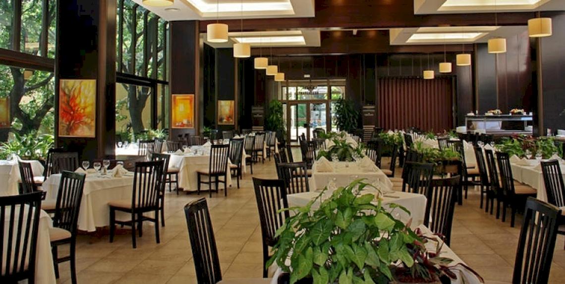 Ein großer Speisesaal im Riviera Beach Hotel mit vielen gedeckten Tischen, Pflanzen und Kunstwerken an den Wänden.
