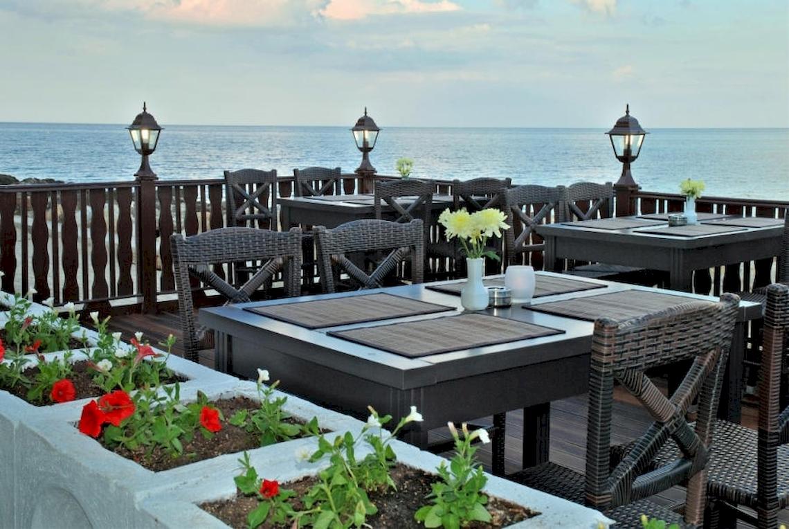 Terrasse des Riviera Beach Hotels mit Blick auf das Meer und mehreren Tischen und Stühlen, die von Blumenbeeten umgeben sind.