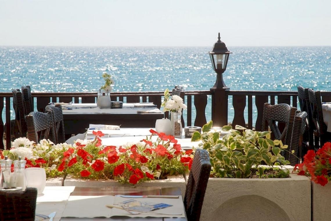 Terrasse mit Meerblick im Riviera Beach Hotel mit blumengeschmückten Tischen und einem Blick auf das offene Meer.