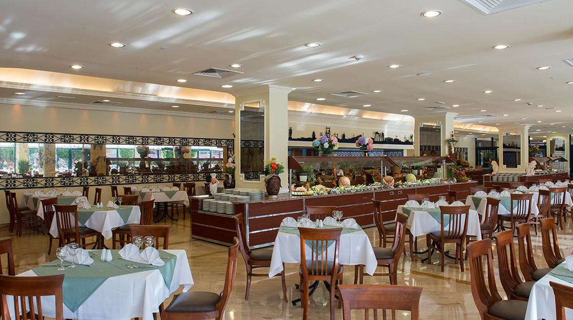 Speisesaal des Miramare Queen Hotels mit vielen Tischen und Stühlen, einer großen Essenstheke und dekorativen Elementen.