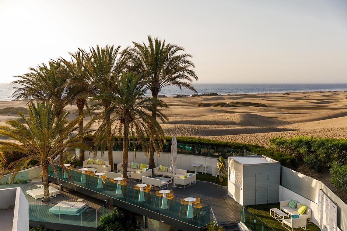 Terrasse des Santa Monica Suites Hotels mit Palmen, weißen Möbeln und Blick auf das Meer und die Dünen.