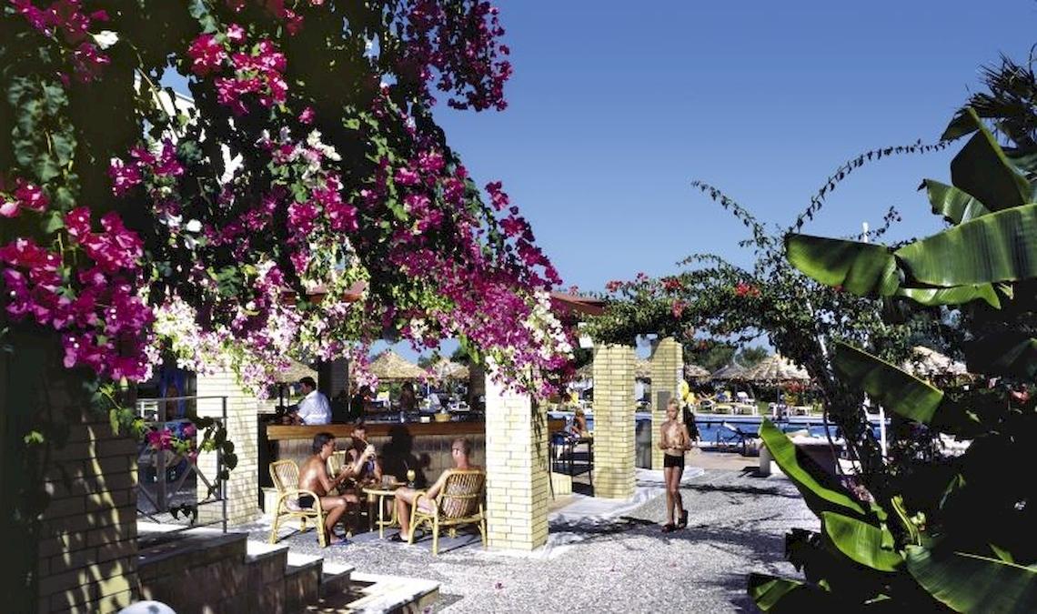 Gäste sitzen draußen an der Bar des Blue Sea Beach Resorts, umgeben von Blumen und Pflanzen.