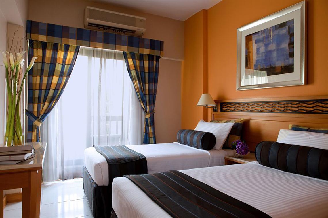 Zwei Betten im Zimmer des Golden Sands Hotel Apartments mit oranger Wand und gemusterten Vorhängen.