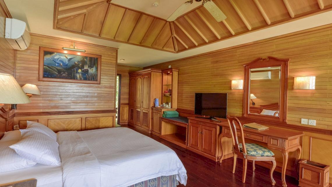 Innenansicht eines Hotelzimmers im Royal Island Resort & Spa mit Holzwänden, einem Bett, Fernseher, Schreibtisch und Möbeln.