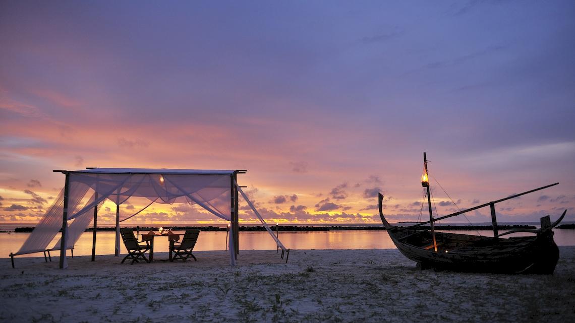 Strandromantisches Dinner unter dem Himmel mit einem Tisch und Stühlen unter einem Baldachin und einem Boot am Ufer.