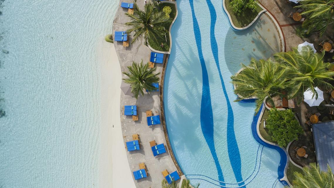 Luftaufnahme des Pools im Royal Island Resort & Spa mit weißen Liegen und blauem Wasser.