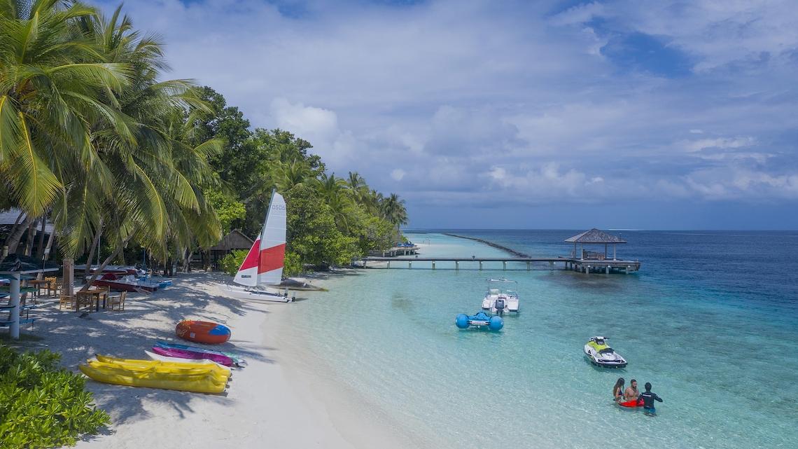 Strandansicht des Royal Island Resort & Spa mit Palmen, Booten und Schwimmern im Wasser.