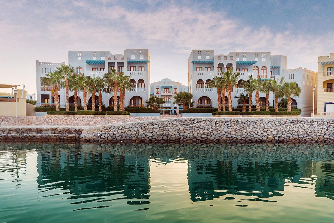 Außenansicht des Fanadir Hotel El Gouna an der Wasserfront mit Gebäuden und Palmen.