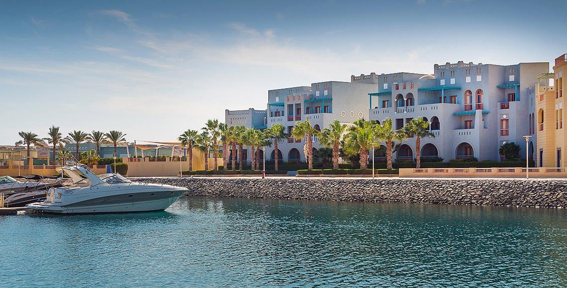 Außenansicht des Fanadir Hotel El Gouna mit weißen Gebäuden, blauen Fenstern und Palmen im Vordergrund.