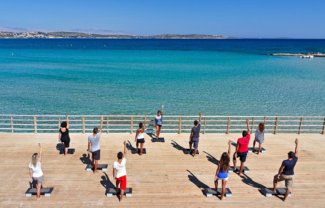 Yoga-Kurs auf Holzplattform am Meer mit Blick auf kristallklares Wasser.