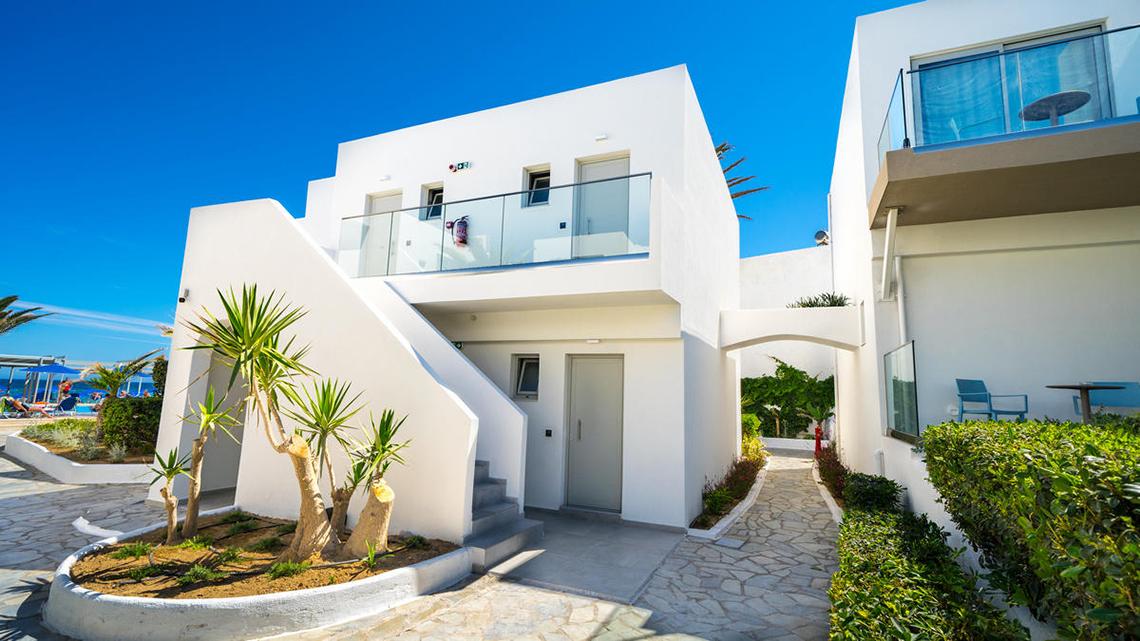 Die Vorderansicht des Adele Beach Hotels zeigt eine weiße Architektur mit Glasbalkonen und einer gepflasterten Einfahrt.