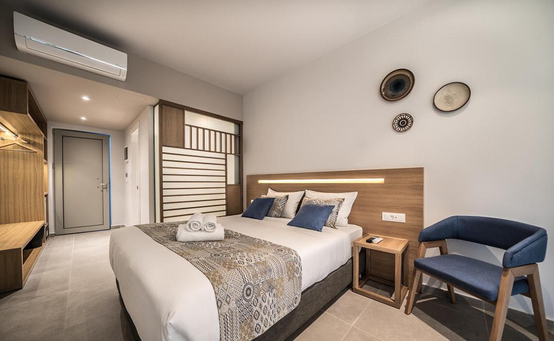 Innenansicht eines Zimmers im Adele Beach Hotel mit Doppelbett, Nachttisch und blauem Stuhl.