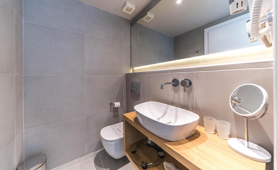 Badezimmer im Adele Beach Hotel mit grauen Fliesen, Toilette, Waschbecken und Spiegeln.