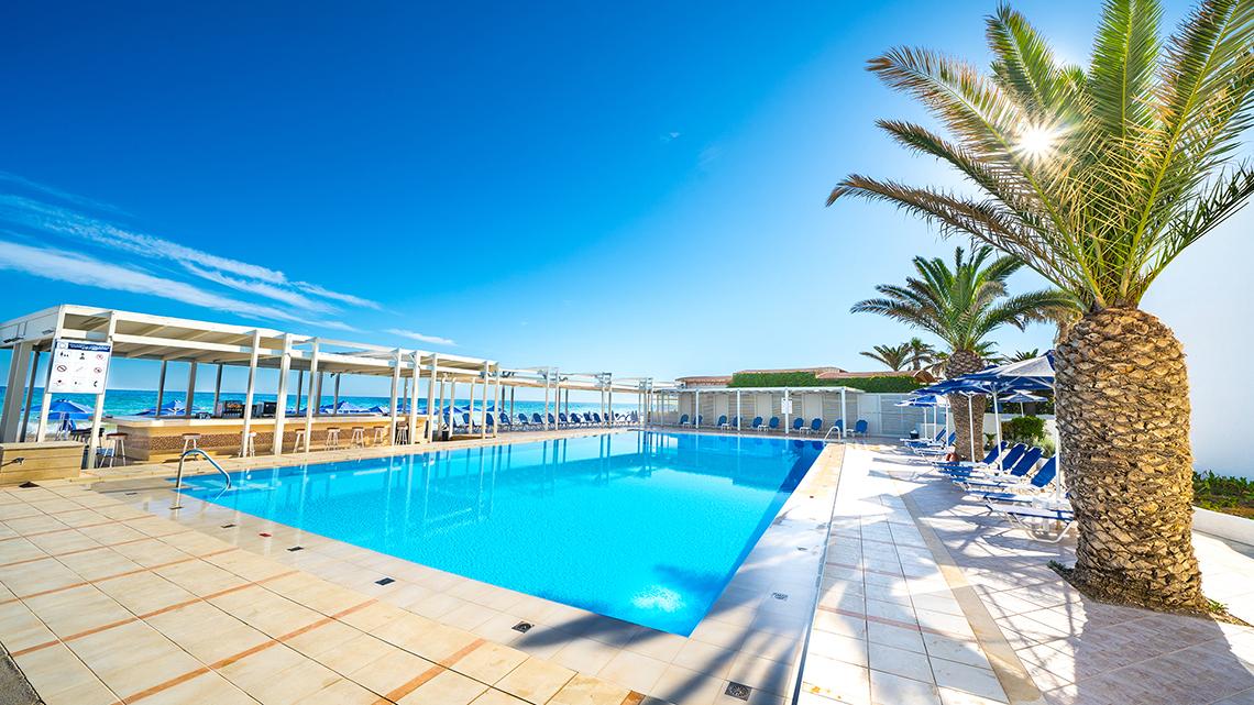 Außenpool des Adele Beach Hotels mit Sonnenliegen und Palmen an einem sonnigen Tag.