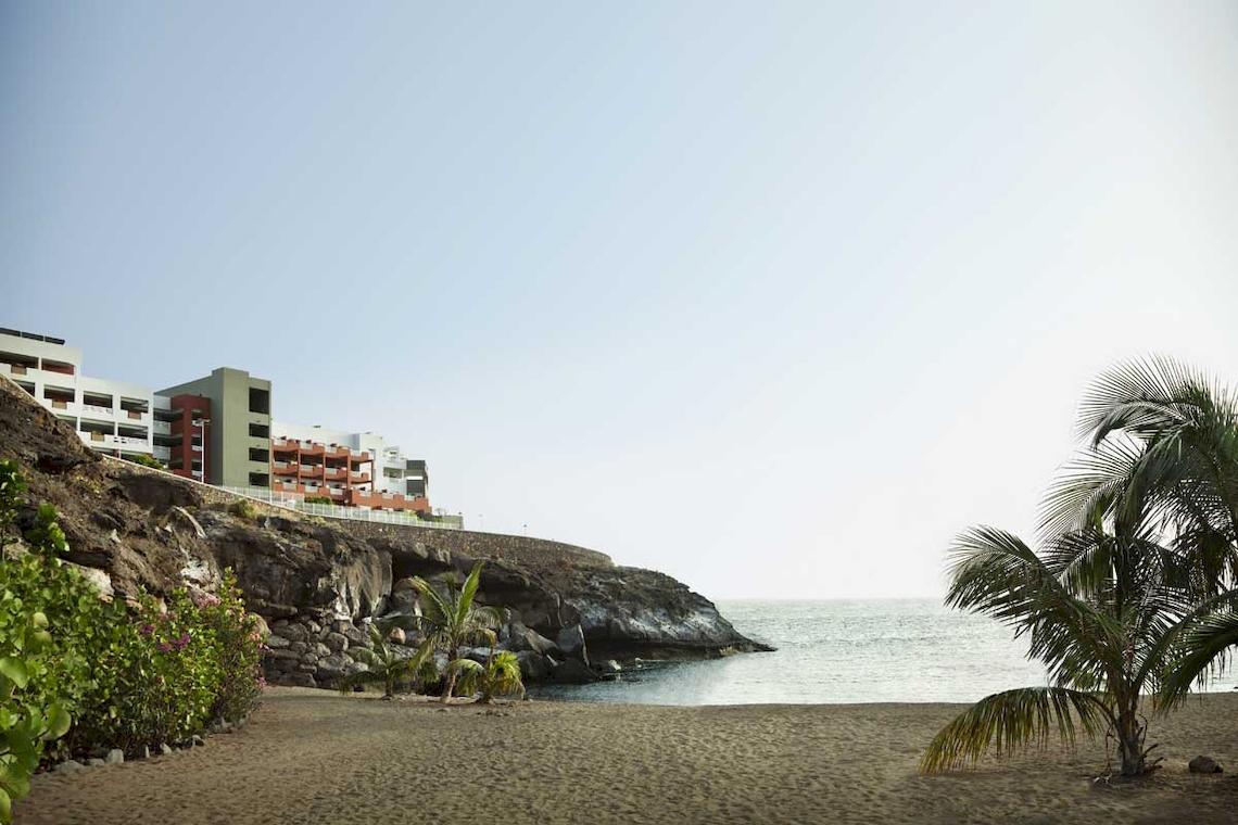 Strand mit Roca Nivaria Gran Hotel im Hintergrund, umgeben von Palmen und Felsen.