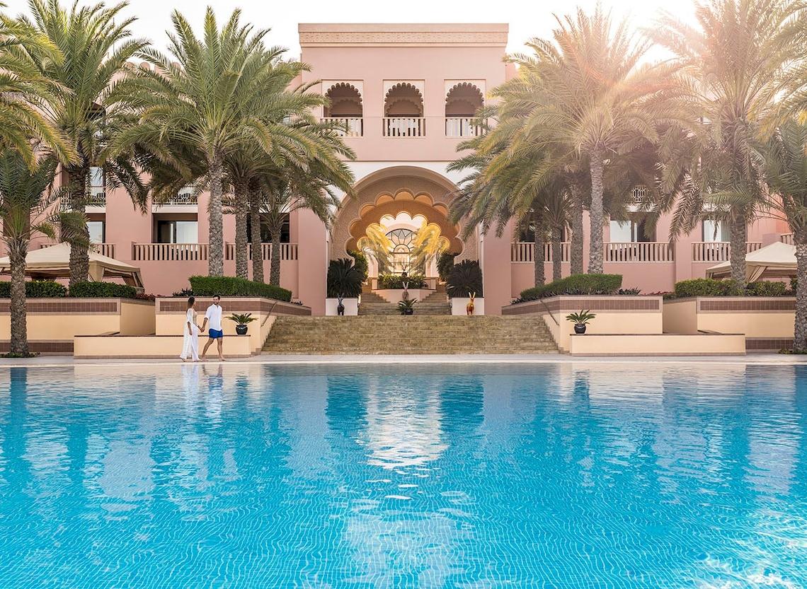 Außenansicht des Shangri-La Al Husn Resort & Spa mit einem großen Pool, Palmen und zwei Personen, die sich unterhalten.