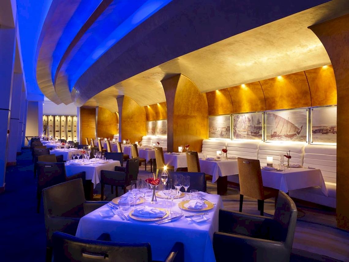 Innenbereich des Shangri-La Al Husn Resorts & Spa, mit blauen Lichtern und goldenen Wänden, gedeckten Tischen und Stühlen.