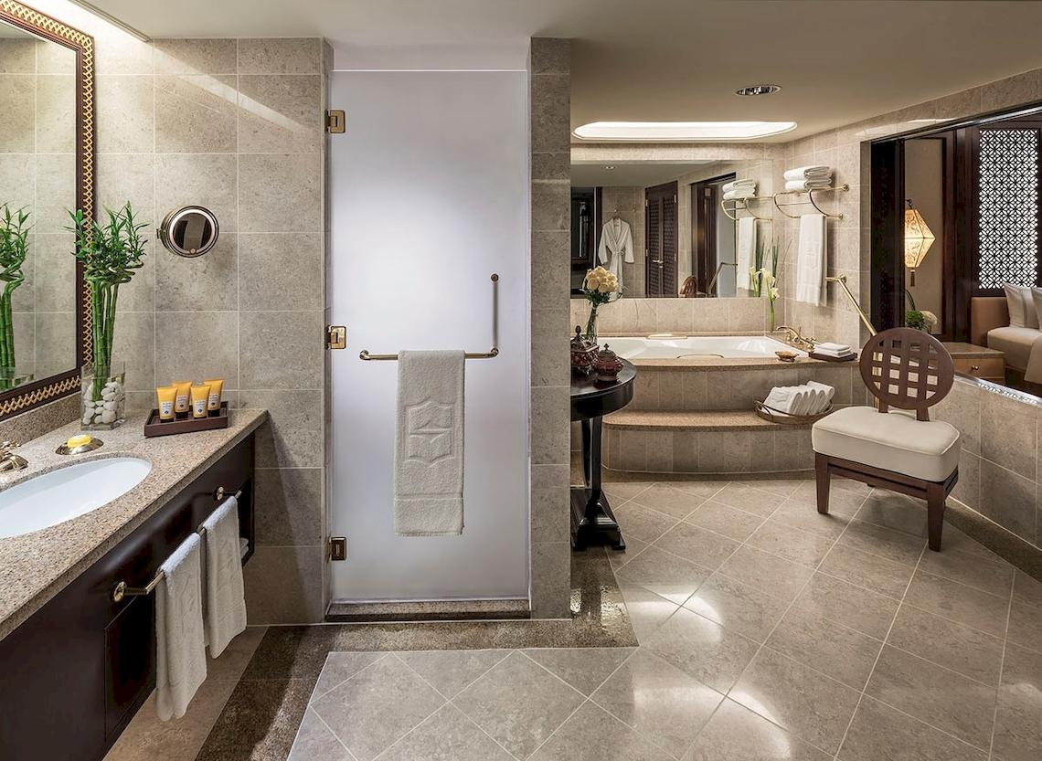 Luxuriöses Badezimmer im Shangri-La Al Husn Resort & Spa mit Badewanne, Dusche, Spiegel und Holzstuhl.