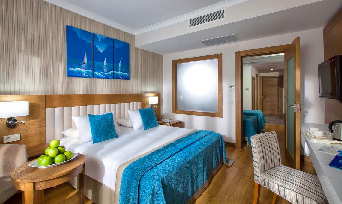 Innenansicht eines Doppelzimmers im Adalya Ocean Deluxe Hotel mit großem Bett, Nachttischen und Kunstwerken an der Wand.