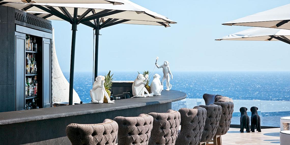 Barbereich des Royal Myconian Resorts mit Blick auf das Meer und weißen Affenstatuen.