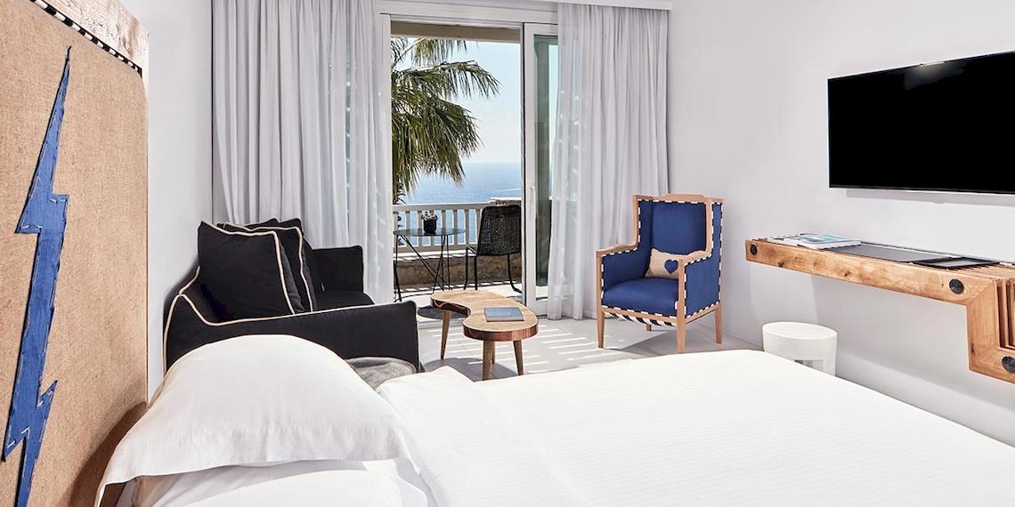Zimmer im Royal Myconian Resort mit Balkon und Meerblick, inklusive Bett, Couch, Fernseher und Nachttisch.