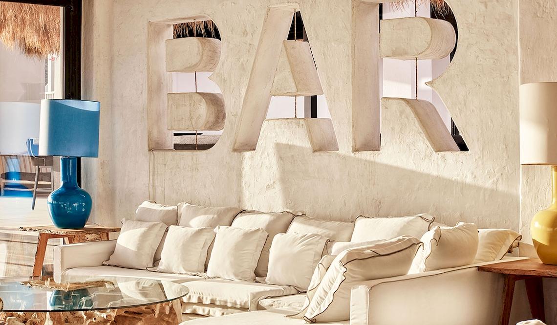 Lounge-Bereich im Royal Myconian Resort mit einem weißen Sofa, Lampen und einer Wand mit dem Wort 'BAR'.