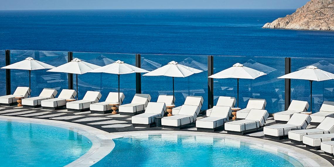 Außenbereich des Royal Myconian Resorts mit Pool, weißen Liegestühlen und Sonnenschirmen vor dem Meer.