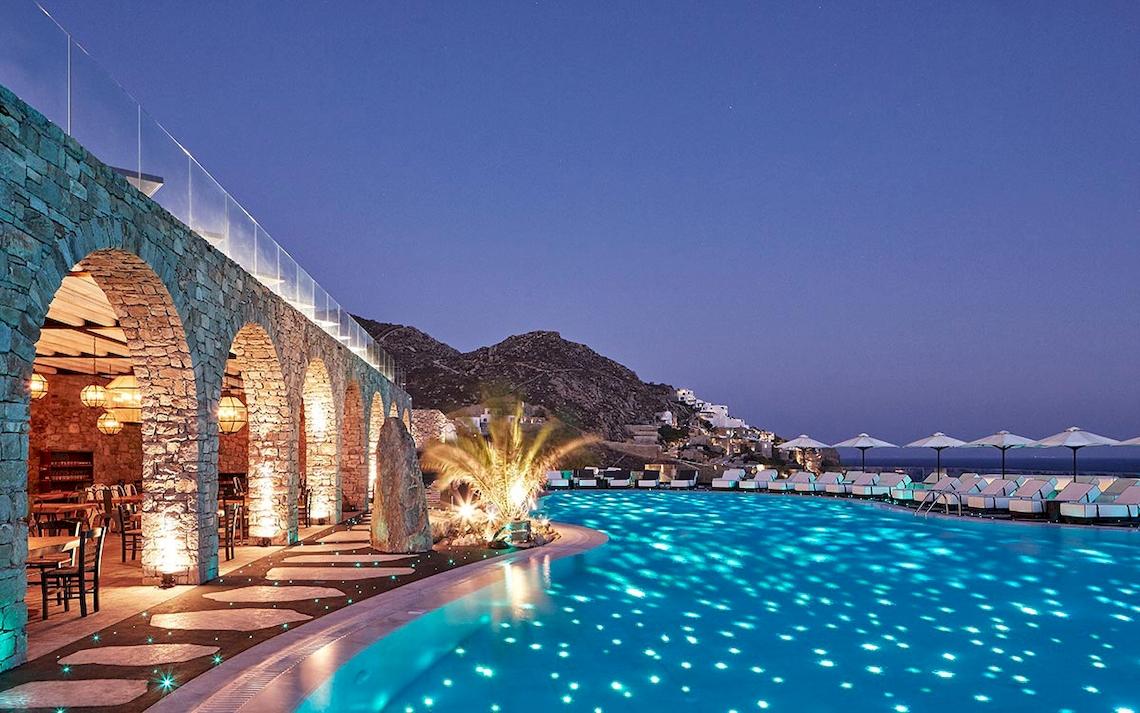 Abendansicht des Royal Myconian Resorts mit beleuchtetem Swimmingpool und Sonnenliegen.