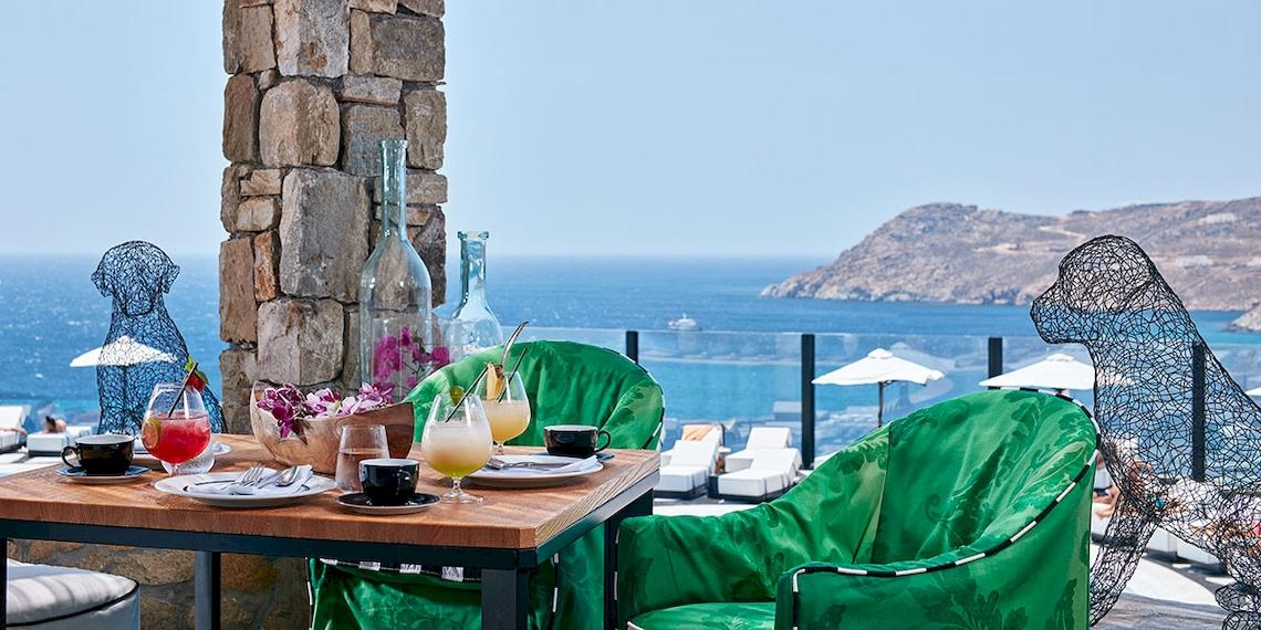 Terrasse des Royal Myconian Resorts mit Blick auf das Meer und grünen Stühlen.