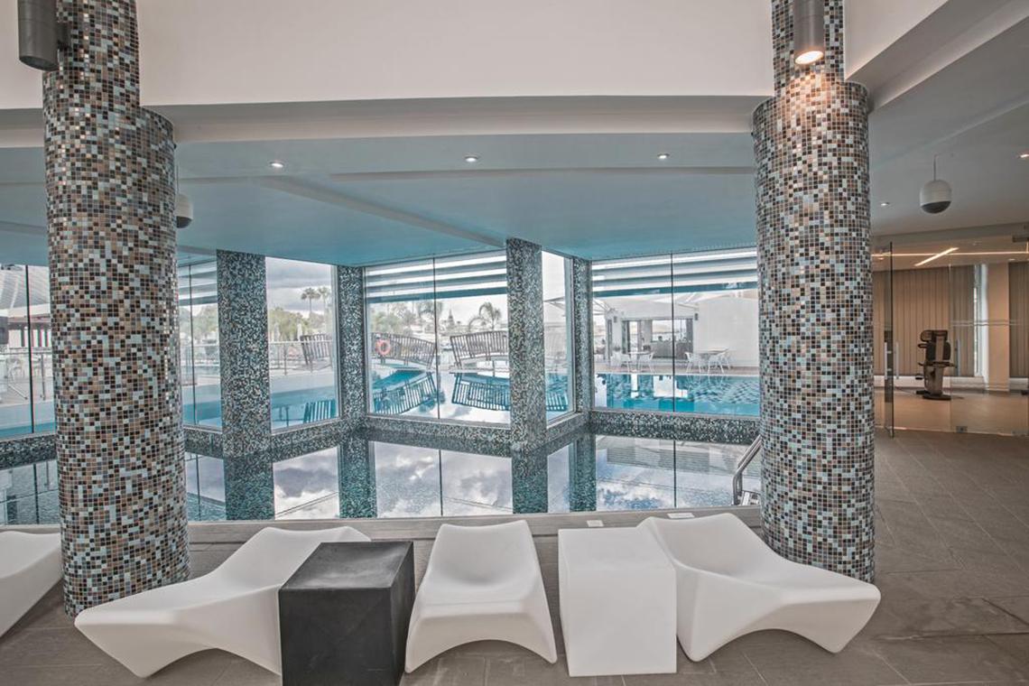 Innenpool-Ansicht im Vassos Nissi Plage Hotel mit weißen Sesseln und gefliesten Säulen.