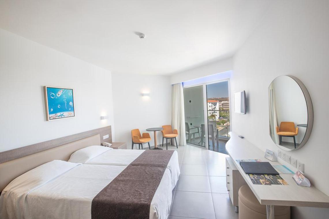Innenansicht eines Zimmers im Vassos Nissi Plage Hotel mit zwei Betten, einem Schreibtisch und einem Balkon.