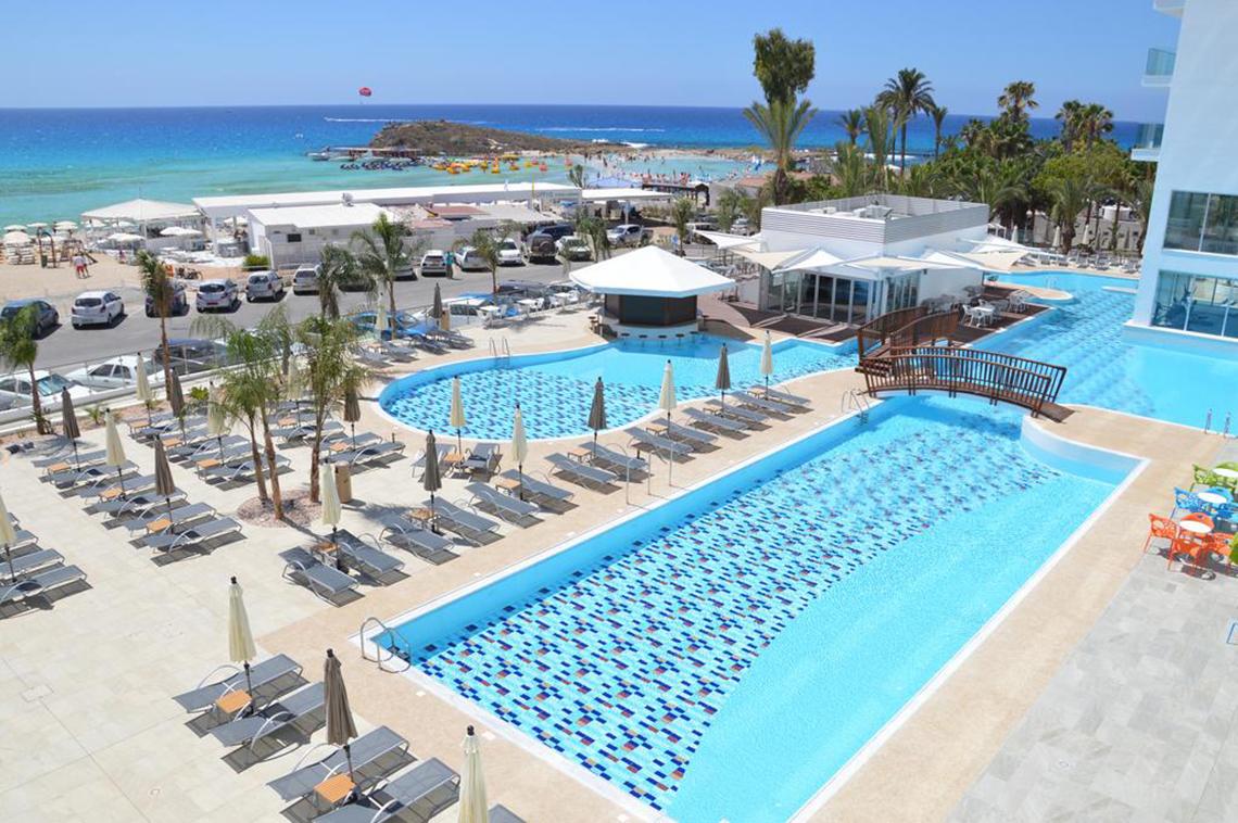 Luftaufnahme des Vassos Nissi Plage Pools mit Sonnenliegen und Meer im Hintergrund