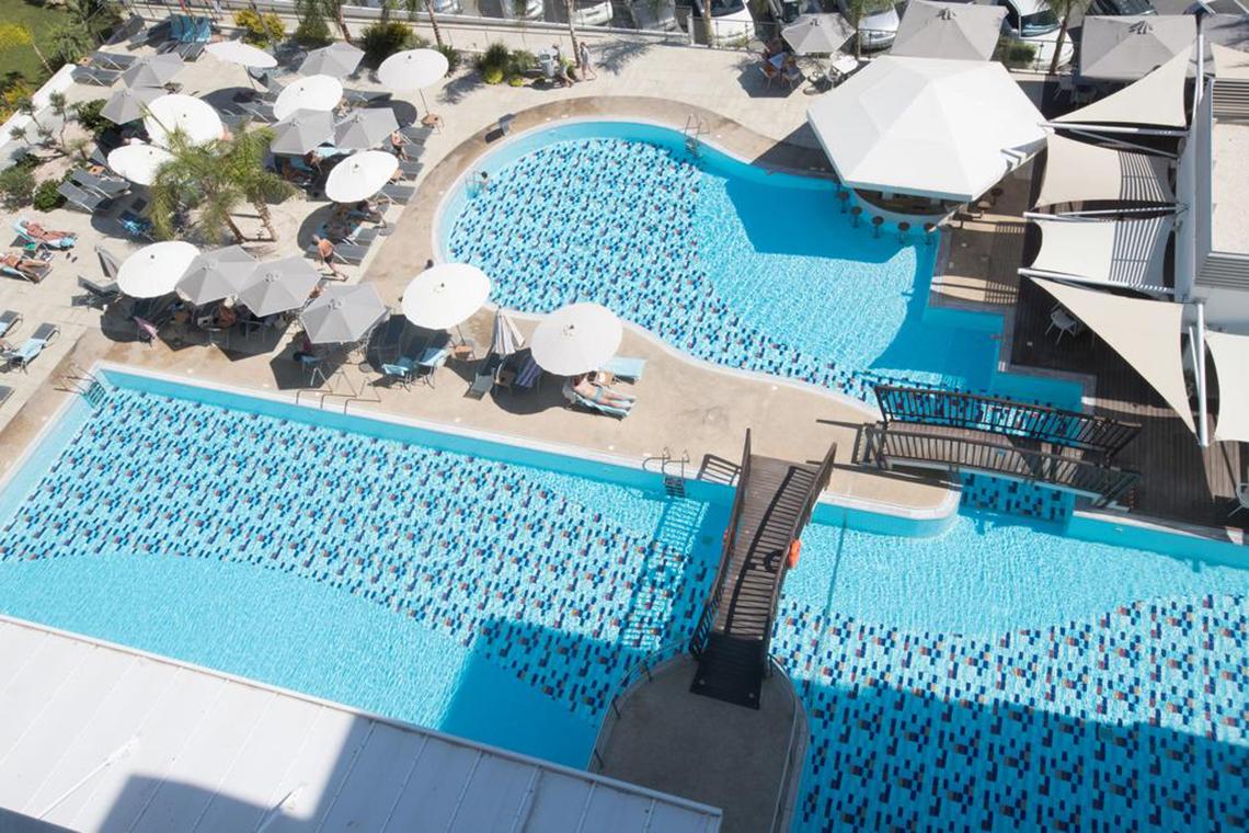 Luftaufnahme des Vassos Nissi Plage Pools mit zwei Becken, Sonnenliegen und Sonnenschirmen.