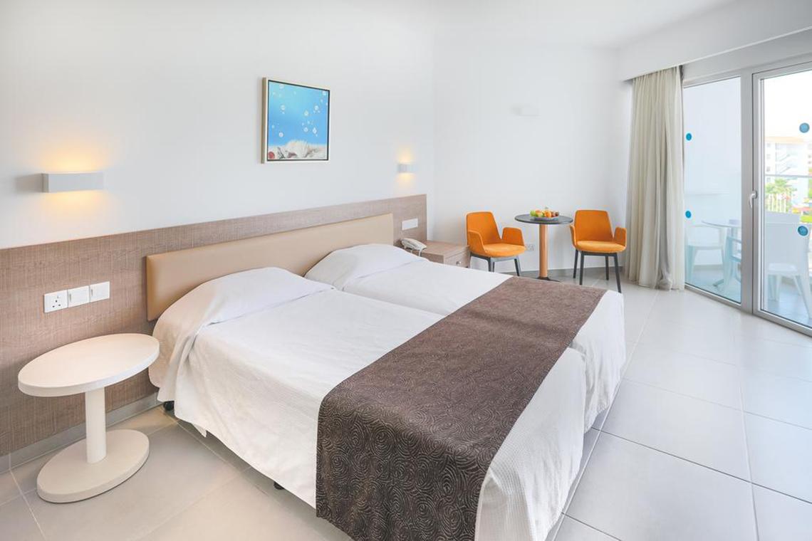 Ein modernes Hotelzimmer im Vassos Nissi Plage mit zwei Betten, einem Tisch und Stühlen vor einer Glastür.