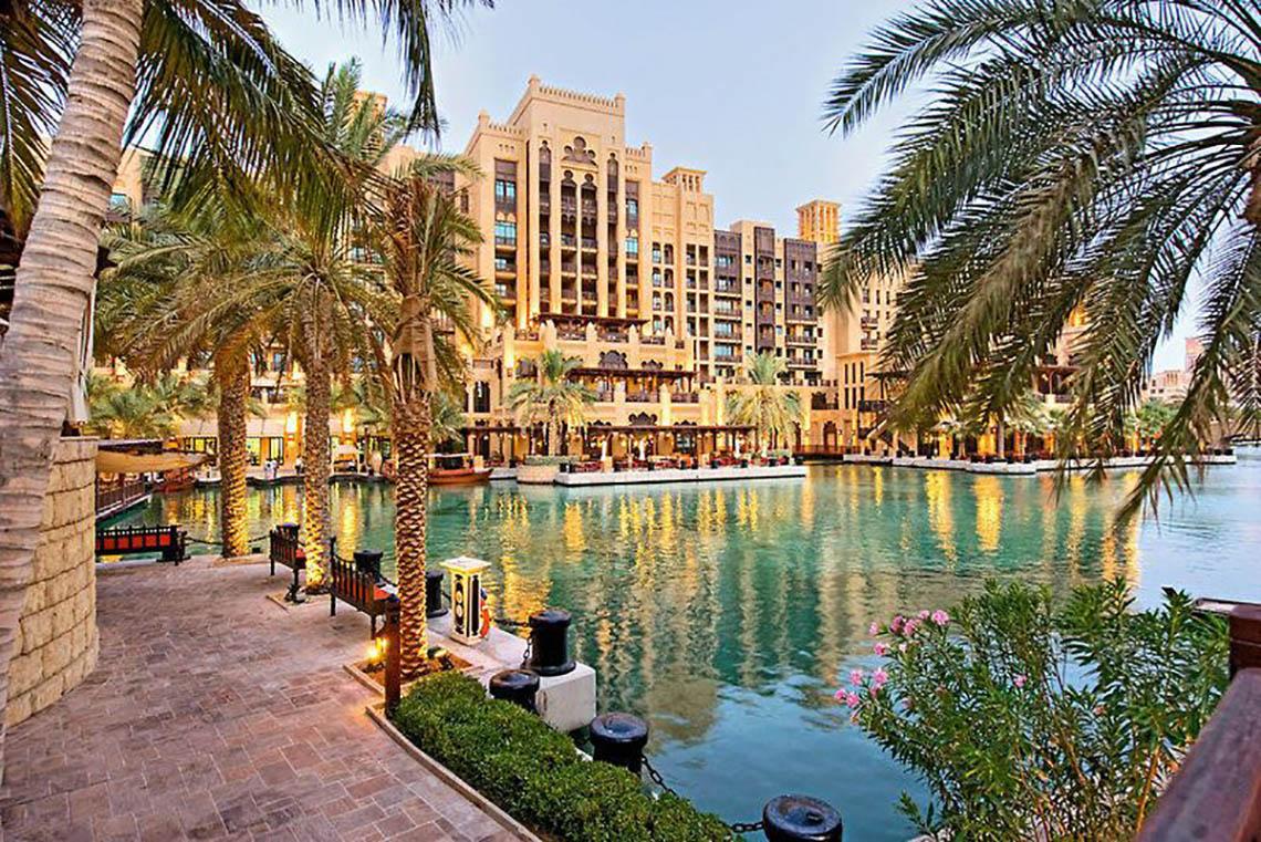 Außenansicht des Jumeirah Dar Al Masyaf Hotels am Wasser mit Palmen und Gebäuden