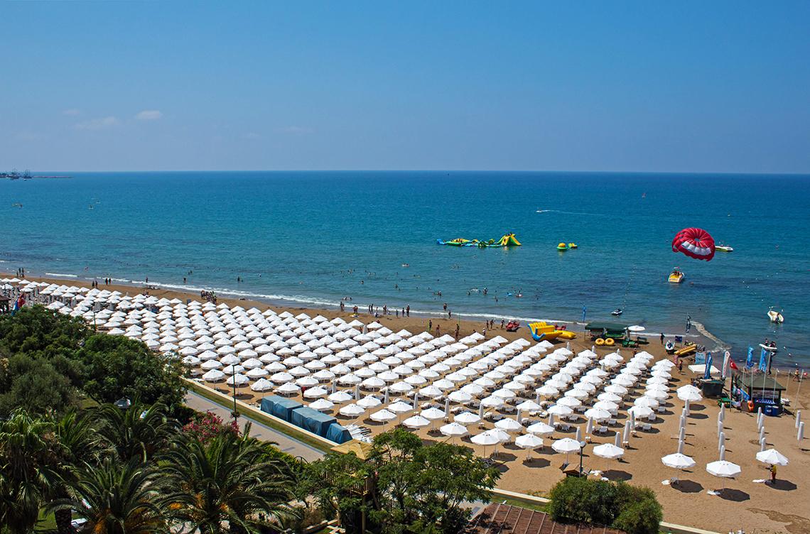 Luftaufnahme des Crystal Sunset Luxury Resort & Spa Strandes mit vielen Sonnenschirmen und Wassersportgeräten