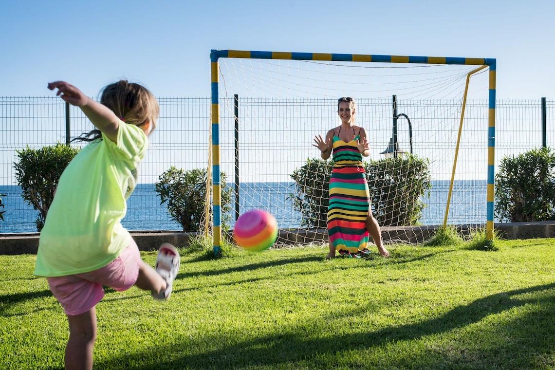 Ein Kind spielt mit seiner Mutter Fußball im Freien, ein Tor im Hintergrund.