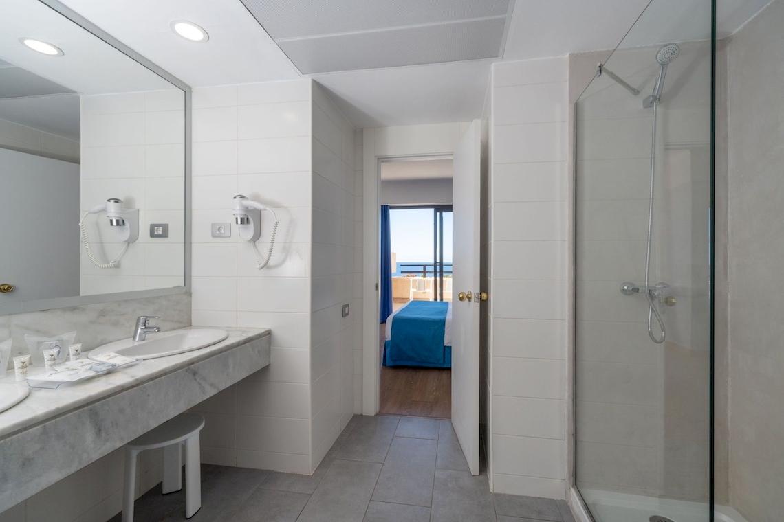 Badezimmer im Grand Teguise Playa mit Doppelwaschbecken, Spiegel, Föhnen und einer offenen Tür zum Schlafzimmer mit einem Bett.