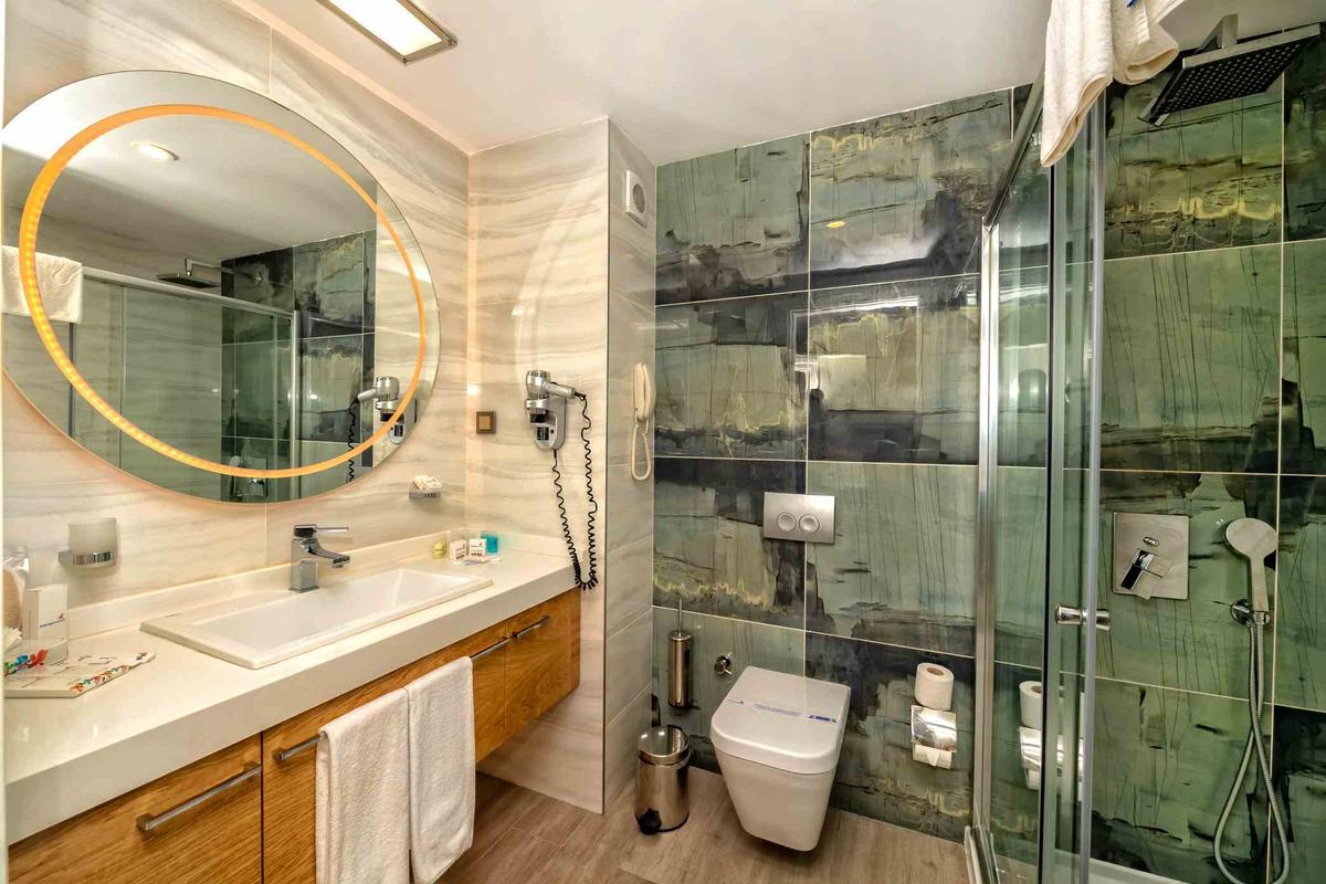 Badezimmer im Sidemarin Kirman Premium Hotel mit ovalem Spiegel, Waschbecken und Duschkabine.