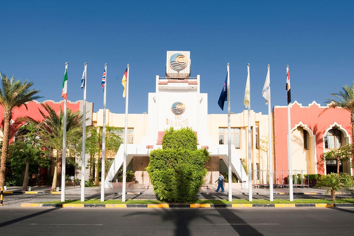 Haupteingang des Grand Hotel Hurghada mit mehreren Flaggen und Bäumen davor.