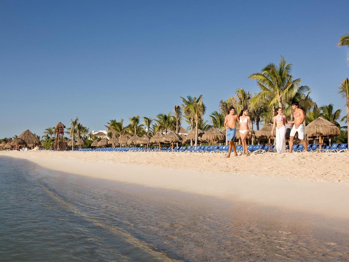 Strand mit mehreren Personen, die auf dem weißen Sandstrand des Grand Sunset Princess All Suites Resorts spazieren.