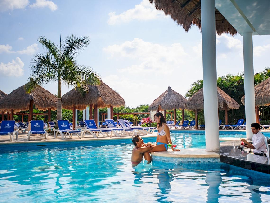 Paar am Poolrand im Grand Sunset Princess All Suites Resort, mit Barkeeper im Hintergrund.
