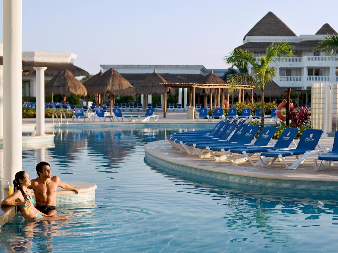 Paar im Pool des Grand Sunset Princess All Suites Resorts mit Liegestühlen und tropischer Umgebung