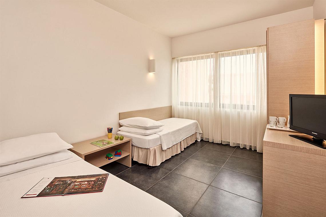 Innenansicht eines Doppelzimmers im Atlantica Mikri Poli Kos Hotel mit zwei Betten, Fernseher und Fenstern.