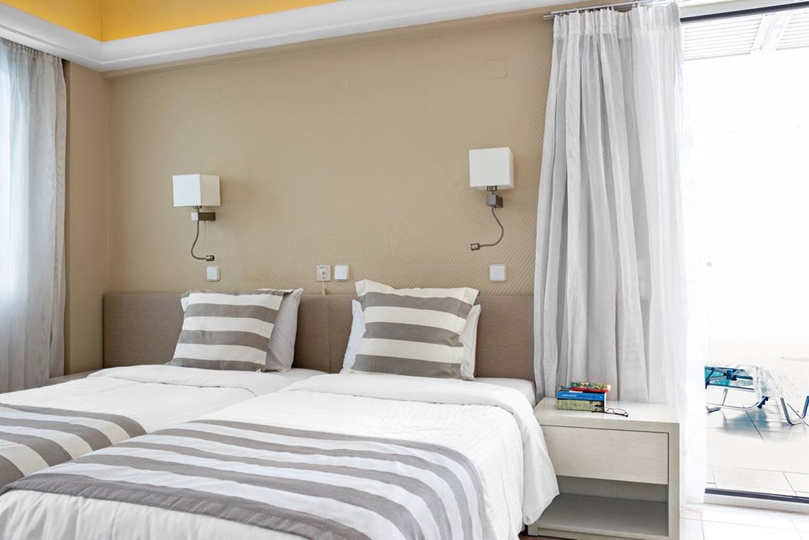 Zwei Betten im Hotelzimmer von Pearle Beach Resort & Spa, beide mit grau-weiß gestreiften Kissen und Decken, neben einem Nachttisch mit Büchern und einer Lesebrille, und einer offenen Tür mit Blick auf einen Balkon.