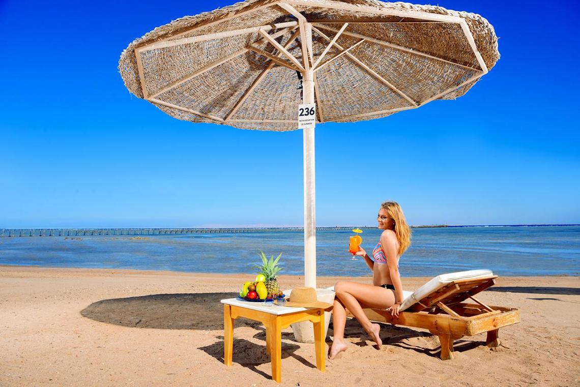 Eine Frau sitzt unter einem Sonnenschirm am Strand und genießt ein Getränk und Obst.