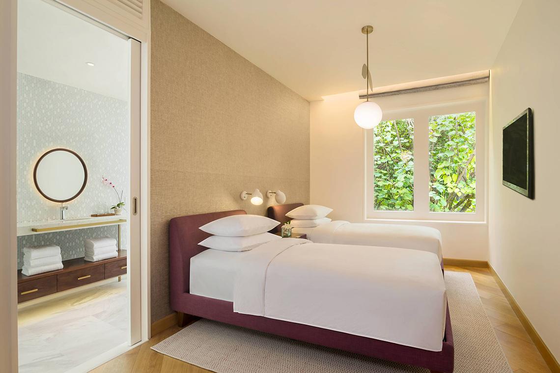 Schlafzimmer im JW Marriott Maldives Resort & Spa mit zwei Betten, weißem Bettwäsche, zwei Lampen und einem Fenster mit Ausblick auf Bäume.