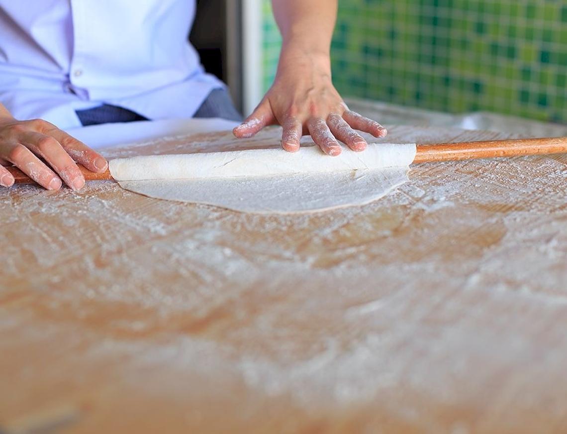 Person rollt Teig mit einer Holzrolle auf einem bemehlten Tisch.