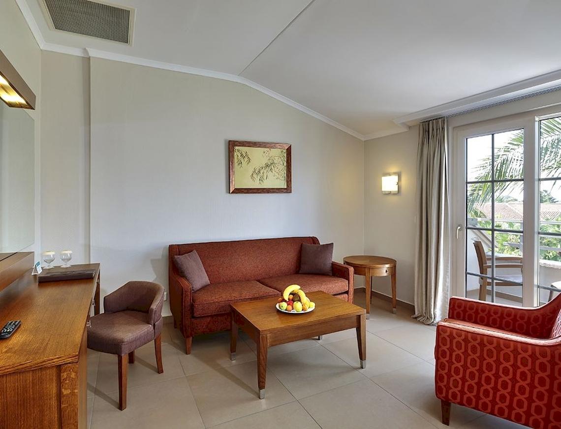 Wohnzimmer des Sunis Elita Beach Resort Hotel & Spa mit rotem Sofa, Tisch, Obstteller und Balkonzugang.